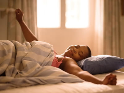 young woman smiling and stretching in bed with early morning light coming in the window behind her